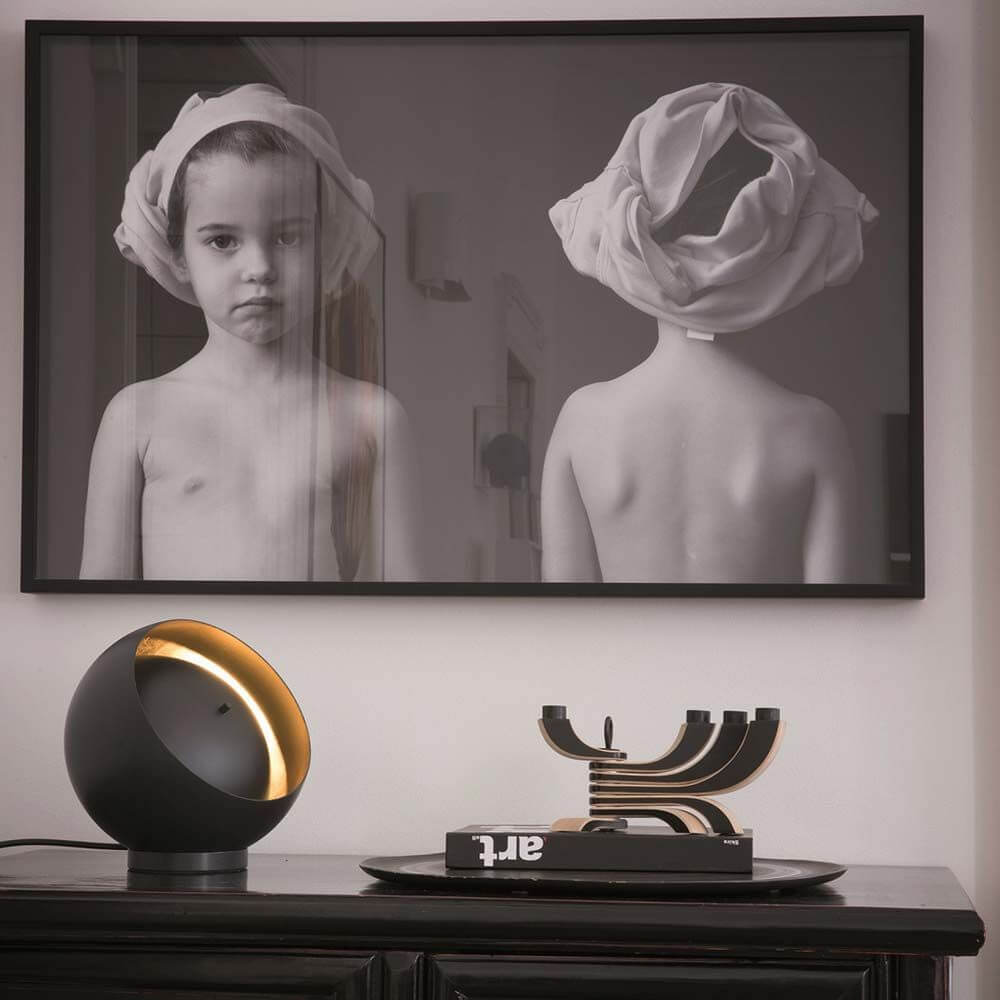 Oluce LED Tischleuchte Eva Schwarz, Goldfarben auf einem Tisch mit einem Kunstwerk im Hintergrund.