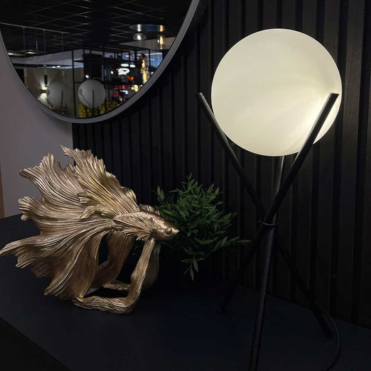 Moderne Tischlampe mit runder Glaskugel, goldener Fischskulptur und grüner Pflanze auf schwarzem Tisch.