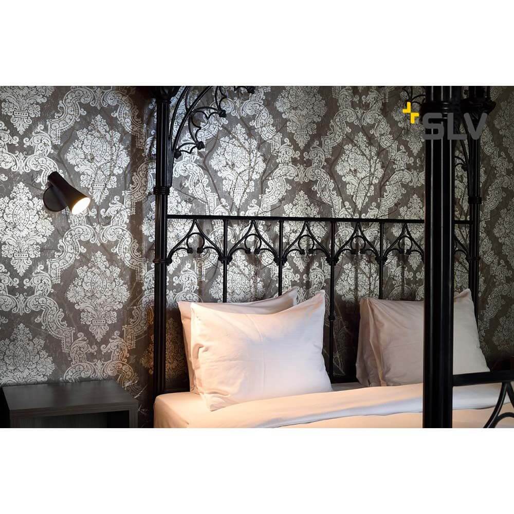 Elegante Phelia Wandleuchte in Schwarz an einem Bett mit edlem Tapetenhintergrund.
