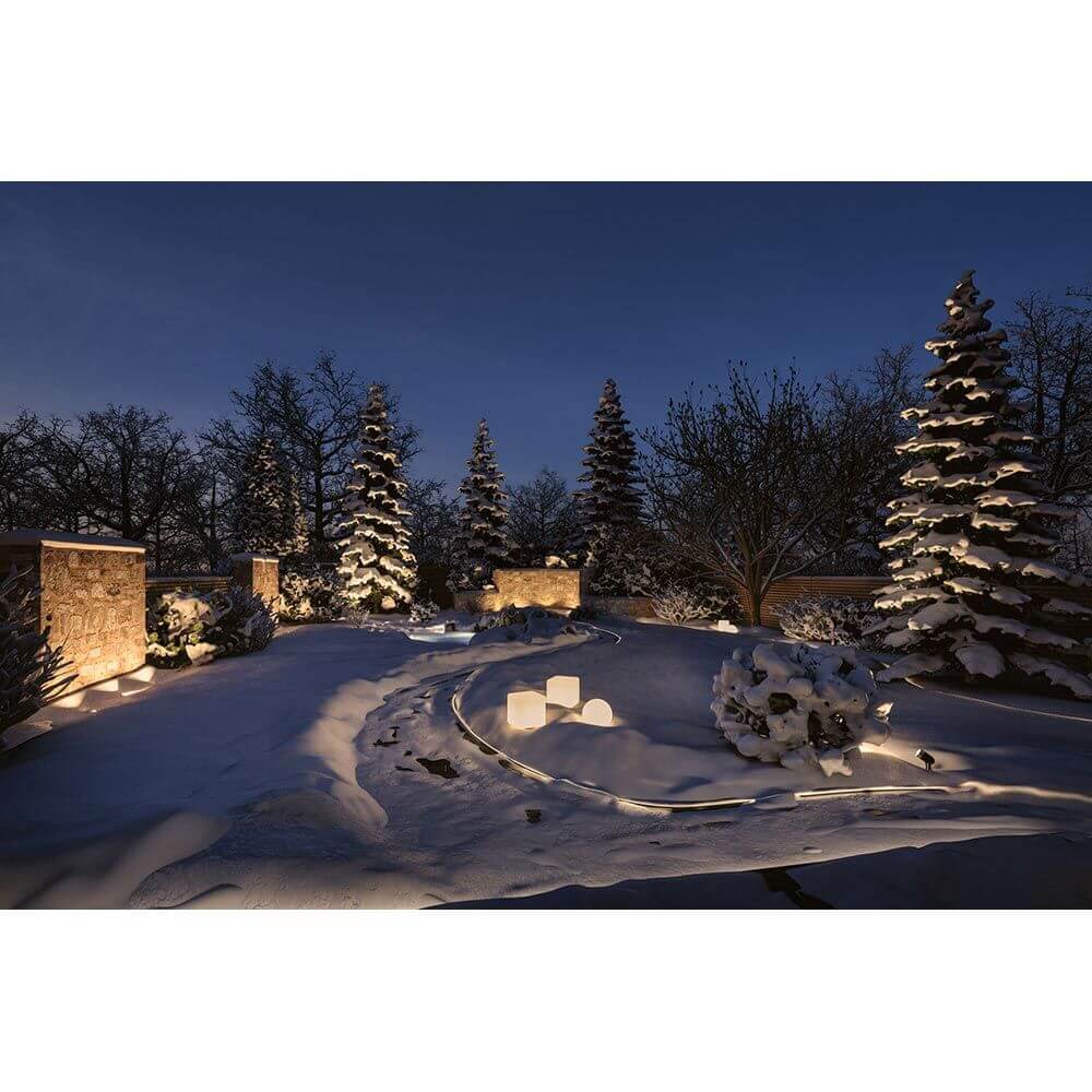 Winterliche Gartenlandschaft mit schneebedecktem Boden und elegantem Lichtdesign zwischen Tannen und Steinen.