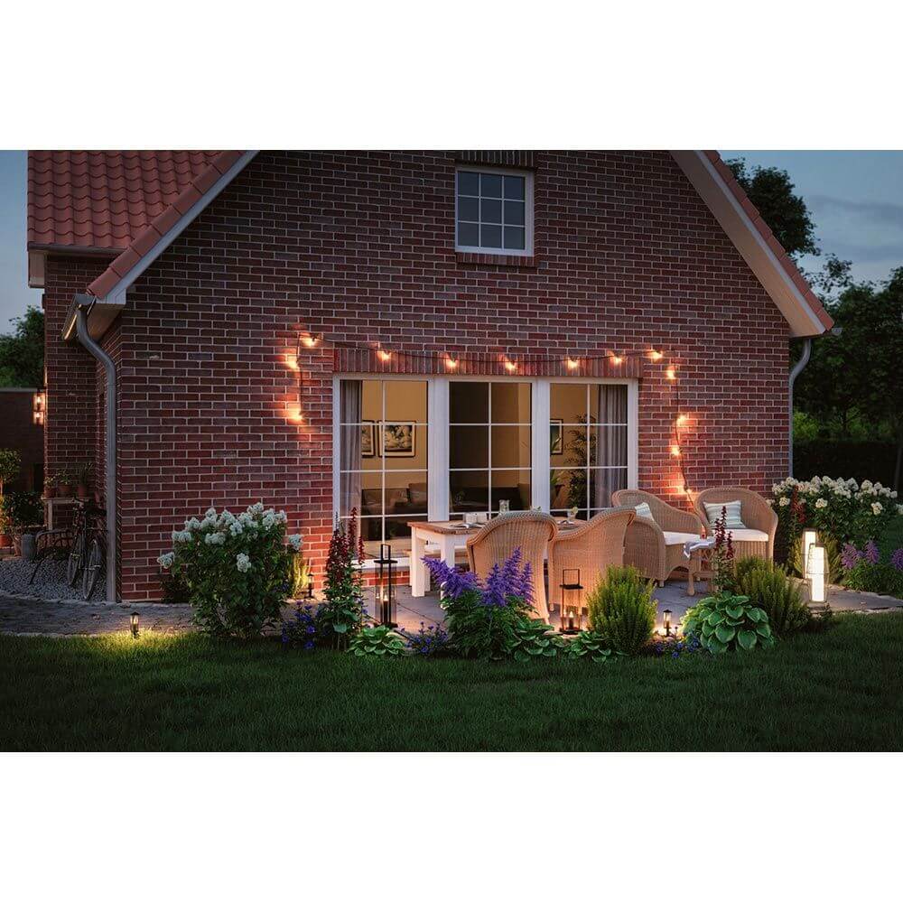 Plug & Shine Classic Laternen Stehleuchte Anthrazit im stimmungsvollen Außenbereich mit Pflanzen und Gartenmöbeln.