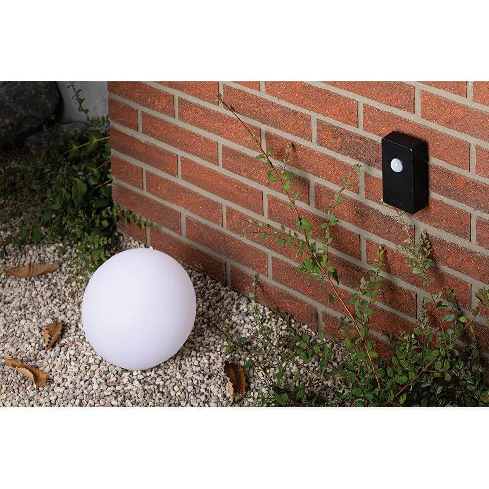 Plug & Shine Smart Home Zigbee Dämmerungssensor Anthrazit neben einer beleuchteten Kugellampe an einer Ziegelwand.