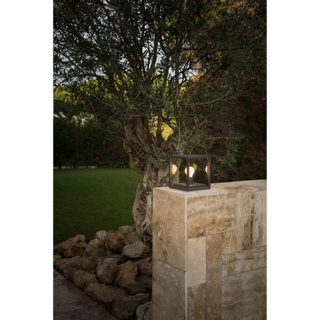 Modern bollard light NALA IP54 dark grey on a stone wall in the garden, illuminated by two light bulbs.