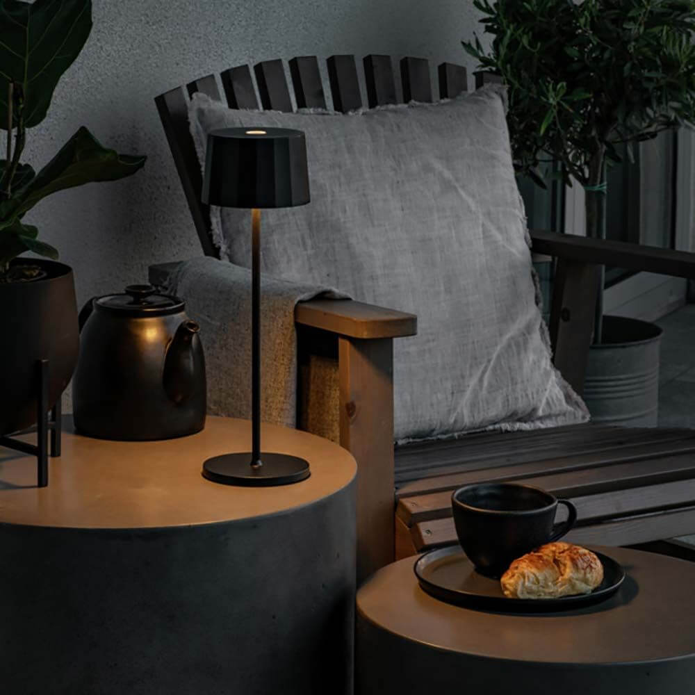 Positano Outdoor LED Akku Tischleuchte auf einem modernen Tisch neben einer Tasse und einem Croissant im Freien.