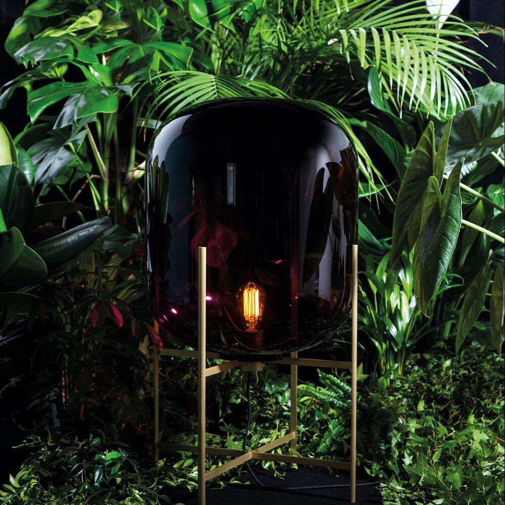 Pulpo LED Tischlampe Oda Medium in schwarzem Glas, von Pflanzen umgeben, industrielles Design mit sanften Rundungen.