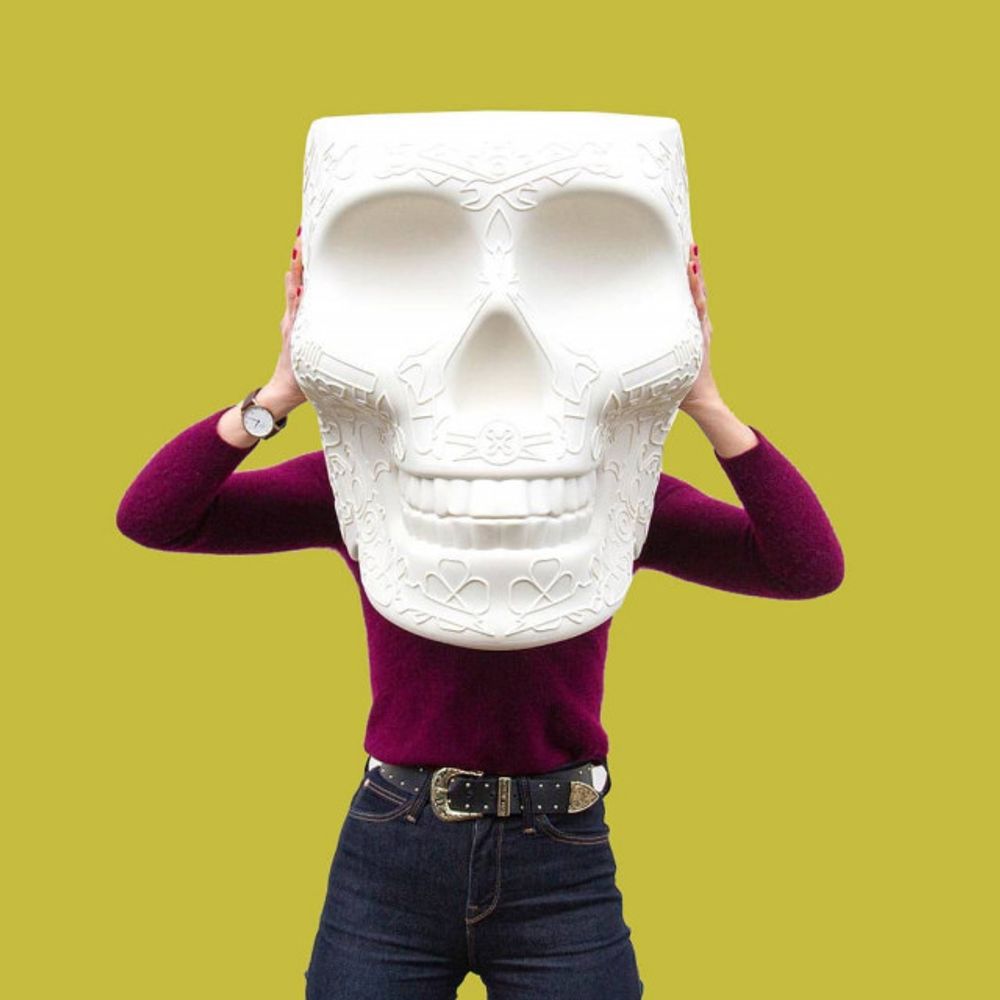 Person hält großen totenschädelartigen Hocker in Weiß vor Gesicht, auf gelbem Hintergrund