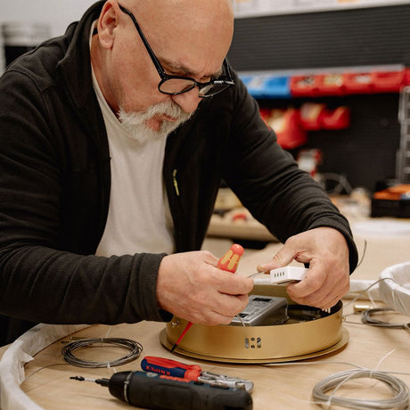 Alt Tag: s.luce Ring Umbau exzentrisch (ohne LED-Ringe) – Handwerker montiert goldenen Ringpendel mit Werkzeug auf Werkbank