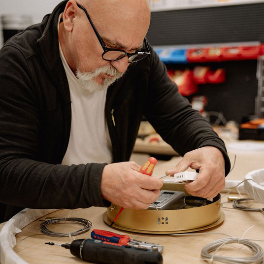 Alt Tag: s.luce Ring Umbau exzentrisch (ohne LED-Ringe) – Handwerker montiert goldenen Ringpendel mit Werkzeug auf Werkbank