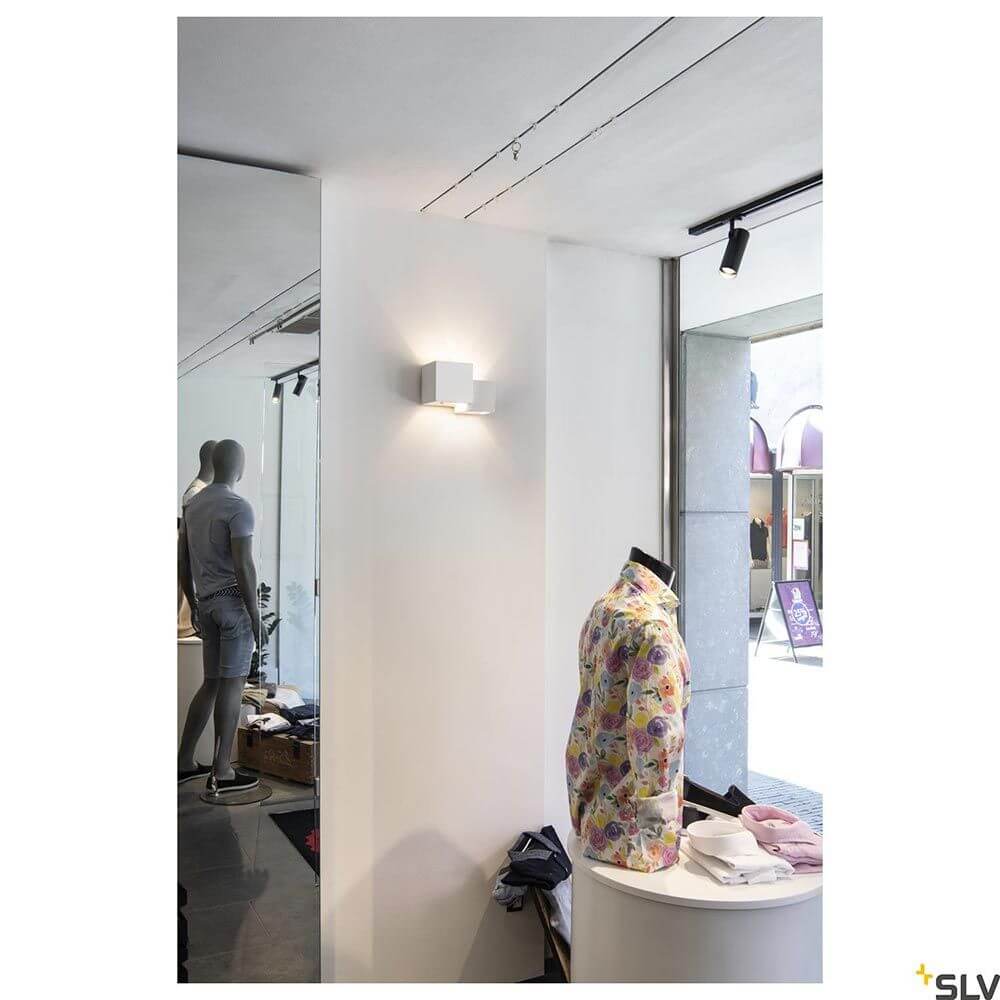 Plastra Cubes Wandleuchte Weiß an einer Wand in einem Modestore, umgeben von Kleidung und Mannequins.
