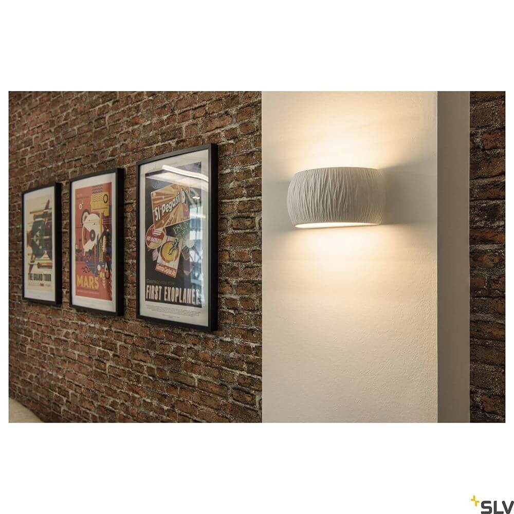 Plastra Curved Wandaufbauleuchte Weiß an einer Steinwand mit gerahmten Bildern im Hintergrund.