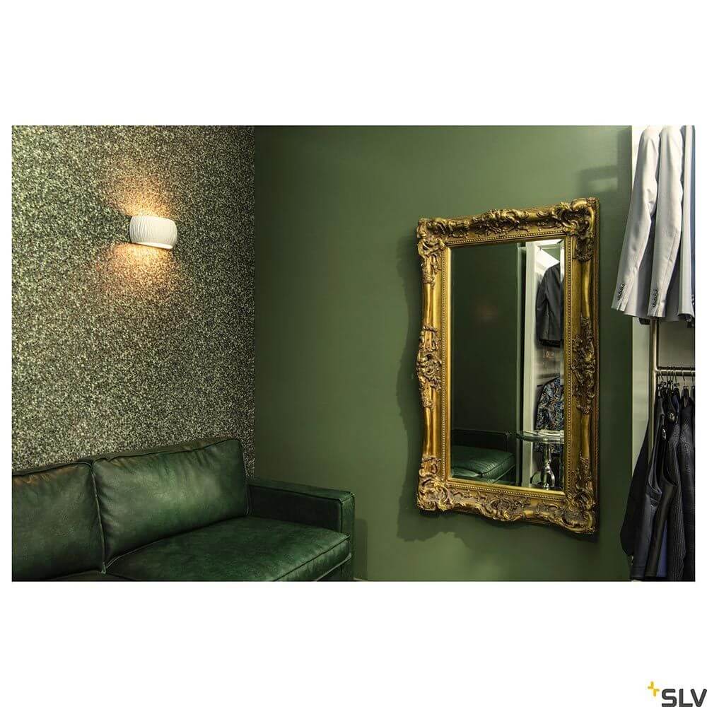 Plastra Curved Wandaufbauleuchte Weiß an einer grünen Wand über einem eleganten Spiegel in einer modernen Einrichtung.