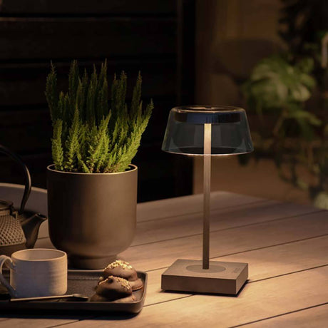 Scilla LED Akku Tischleuchte IP54 auf einem Tisch, umgeben von Pflanzen und einer Tasse, ideal für stimmungsvolle Beleuchtung.