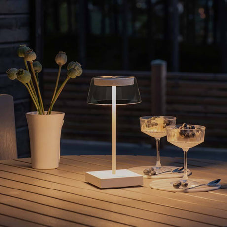 Scilla LED Akku Tischleuchte IP54 auf einem Tisch, umgeben von Pflanzen und Gläsern bei Dämmerung.