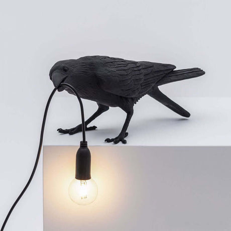 Lampe de table d’extérieur Seletti Bird inclinée, noire, corbeau avec câble et ampoule sur un socle blanc.