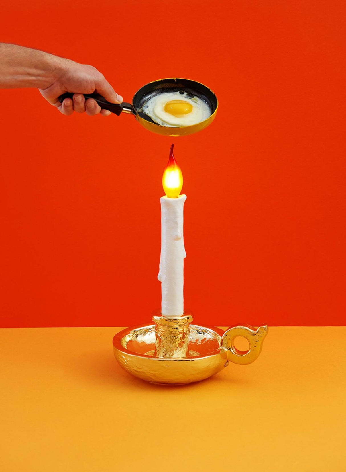 Seletti Grimm Akku-LED-Tischleuchte dimmbar: goldener Kerzenleuchter-Design mit LED-Flamme und Spiegelei-Pfanne vor rotem Hintergrund