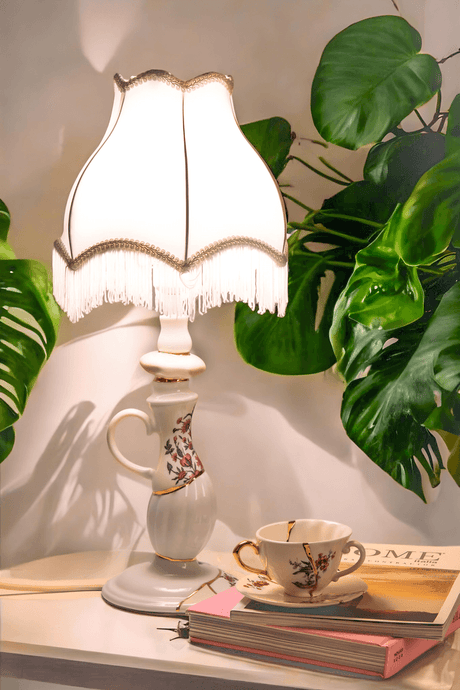 Lampe de table Seletti Kintsugi dimmable en porcelaine avec finition dorée, allumée sur une table de chevet à côté d’une tasse à thé vintage et de plantes