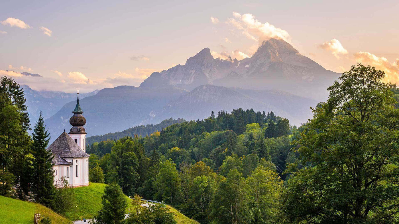 Blick auf eine malerische Kirche in den Bergen, umgeben von grünen Wäldern und majestätischen Gipfeln.