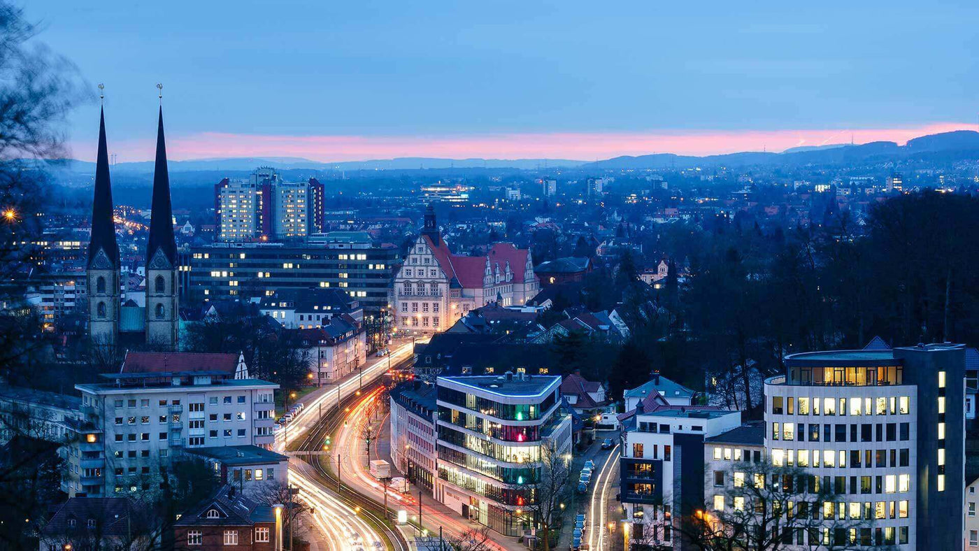 Panorama einer Stadt bei Dämmerung, mit lichterfüllten Straßen und Blick auf historische Gebäude und moderne Architektur.