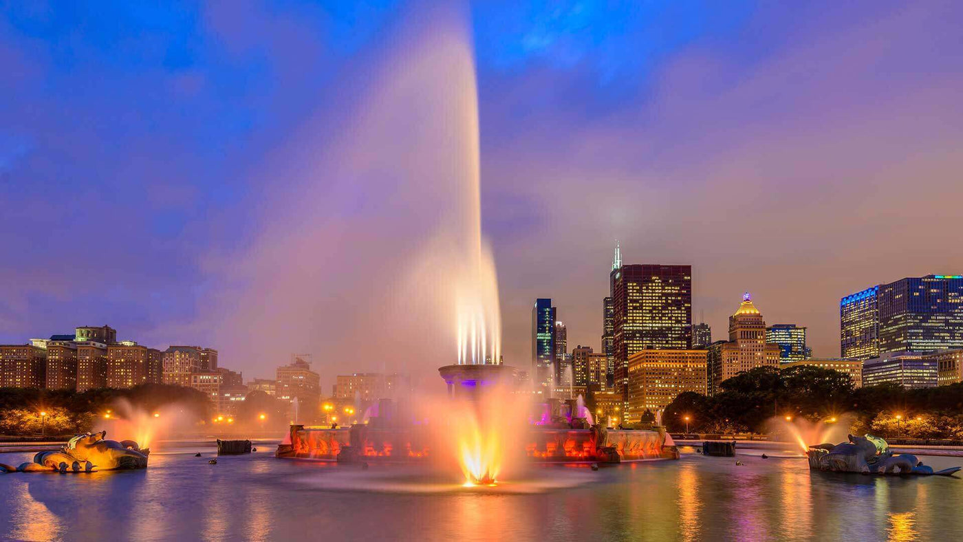 Buckingham Fountain bei Nacht mit beleuchtetem Wasserstrahl und Stadtansicht von Chicago im Hintergrund.
