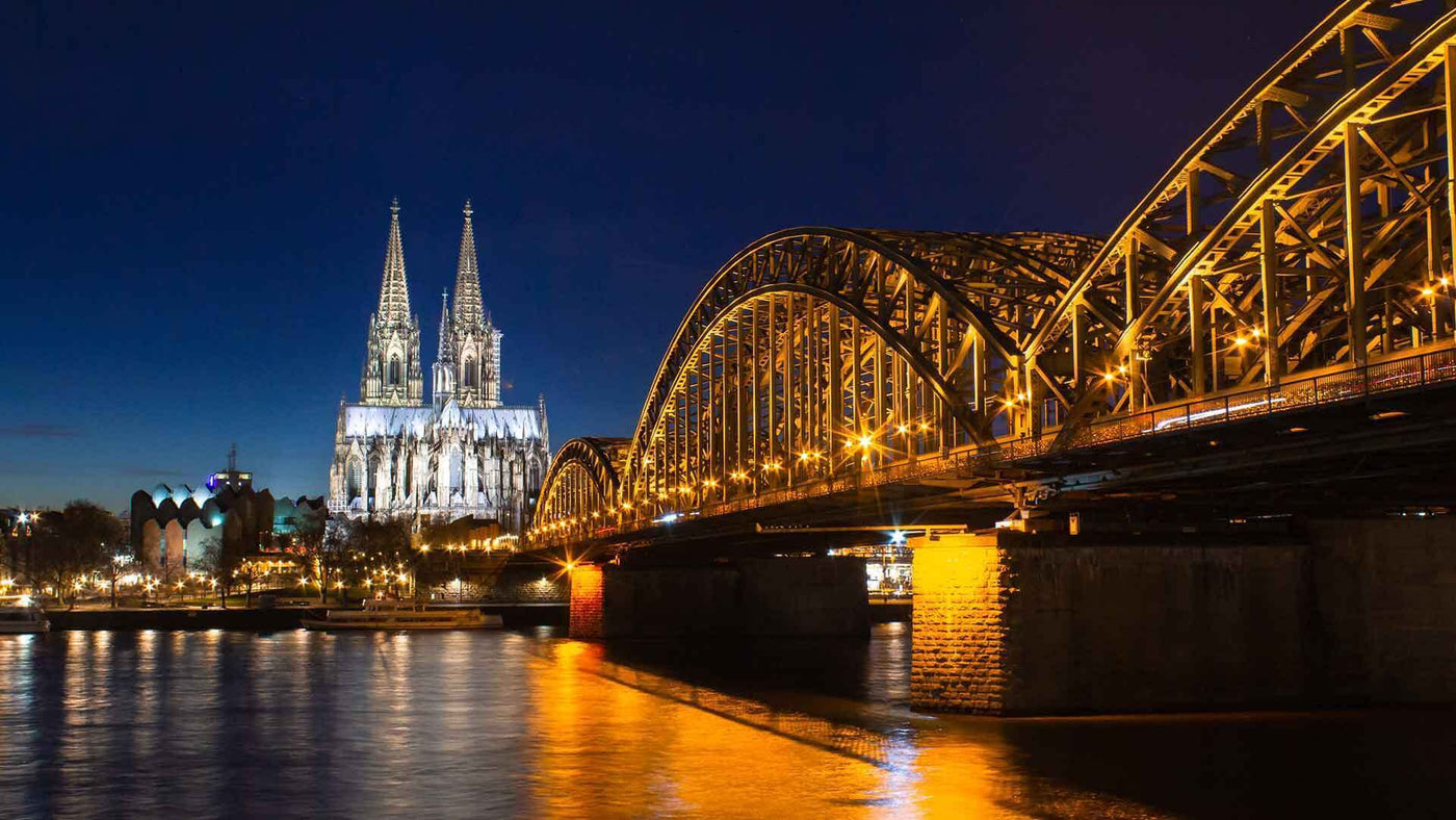 Kölner Dom und Hohenzollernbrücke bei Nacht, erleuchtet im Schatten des Abendhimmels.
