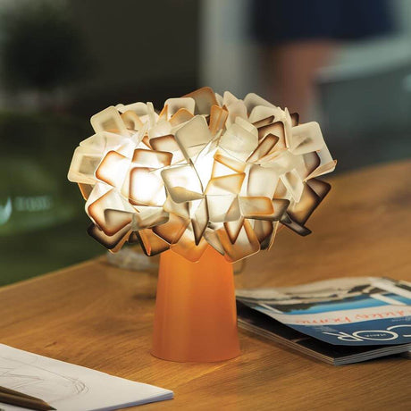 Lampa stołowa Slamp Clizia Orange na stole, o nowoczesnym, kwiatowym designie i ciepłym świetle.