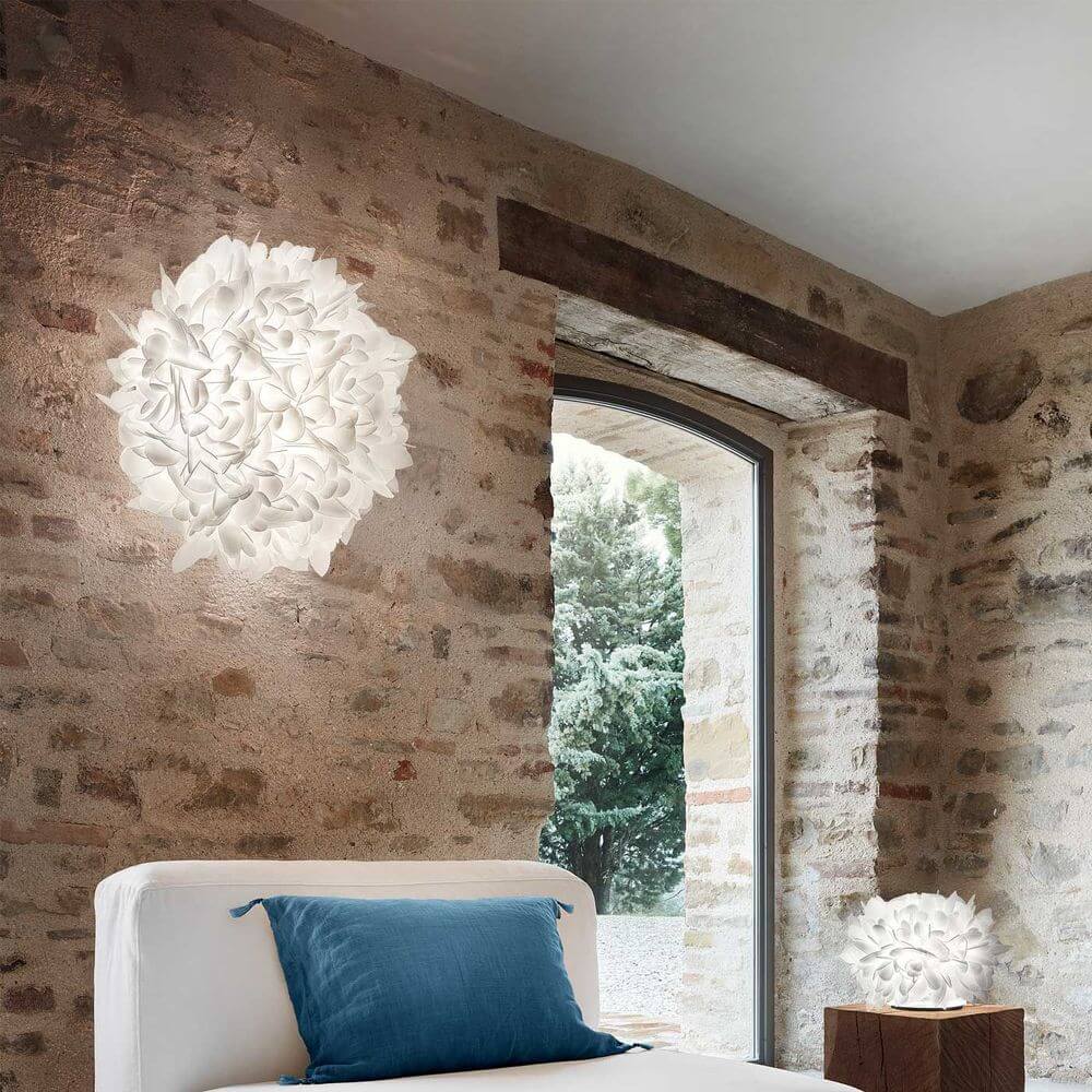 Slamp Wand- & Deckenlampe Veli Foliage an einer Steinwand über einem Bett platziert, mit Fensterblick.