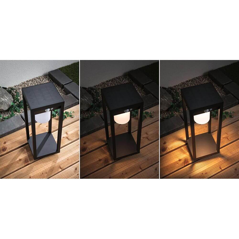 Solar LED Sensor Laterne Marisol Schwarz bei Nacht mit warmweißem Licht auf Holz- und Steinboden.