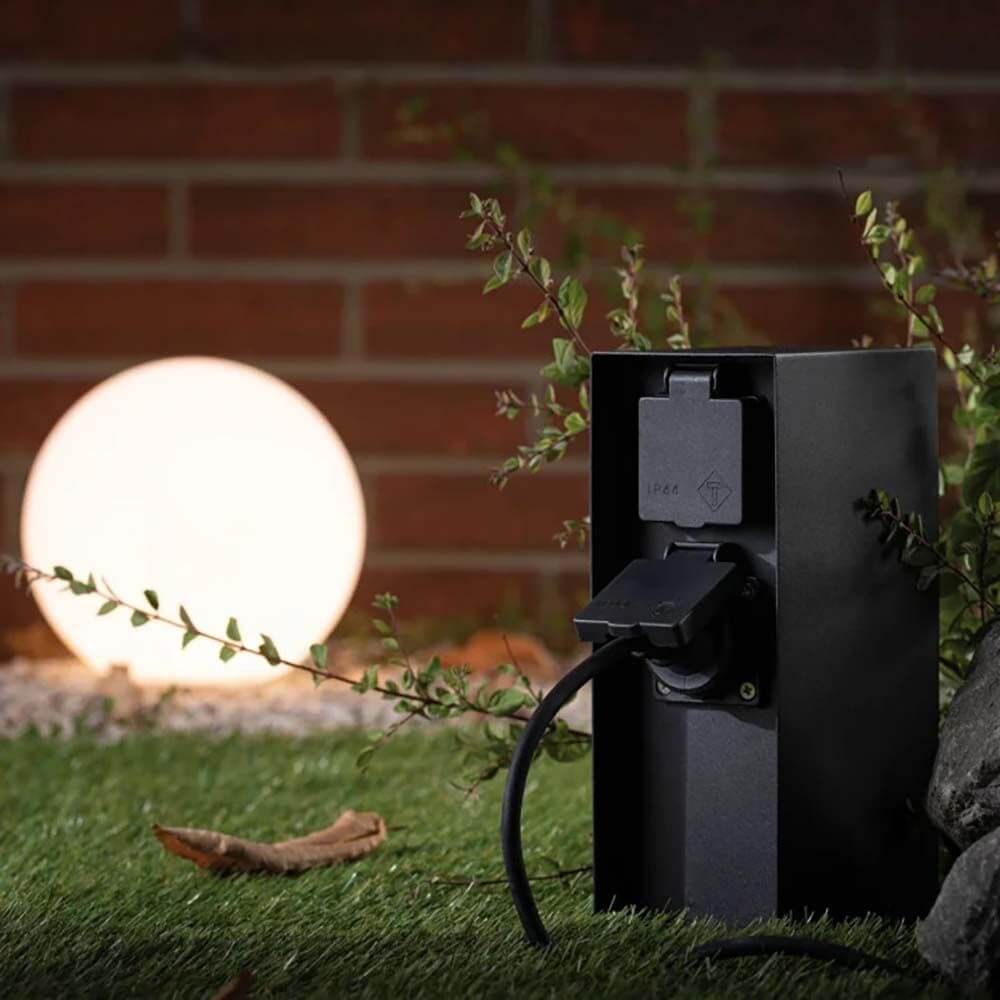 Steckdosensäule Rio IP44 Grau im Garten mit beleuchtetem Kugelleuchten-Hintergrund, ideal für Außenstromeinsatz.