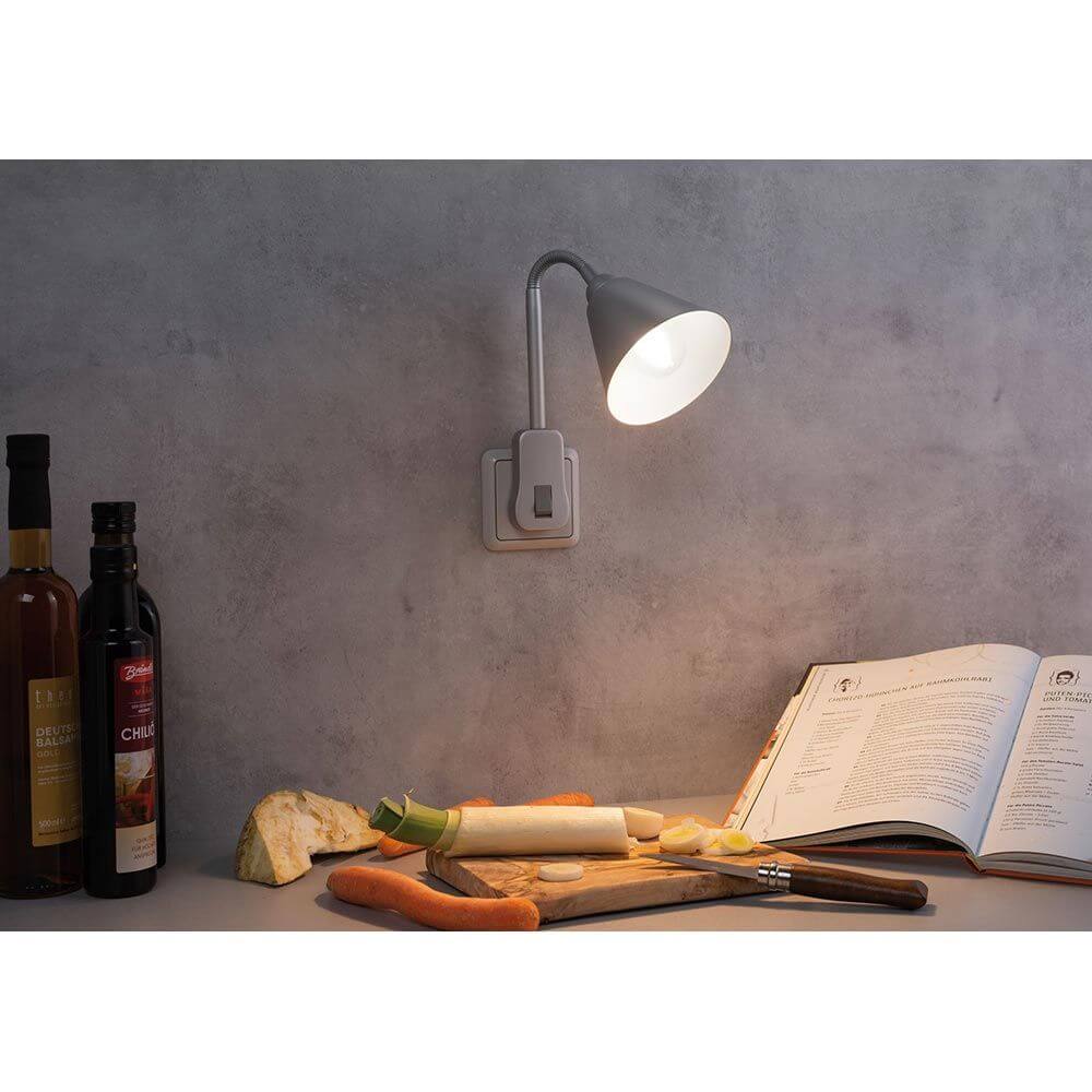 Steckerleuchte Junus Flexarm Grau an einer Wand mit einem Kochbuch und Lebensmitteln auf einem Tisch.