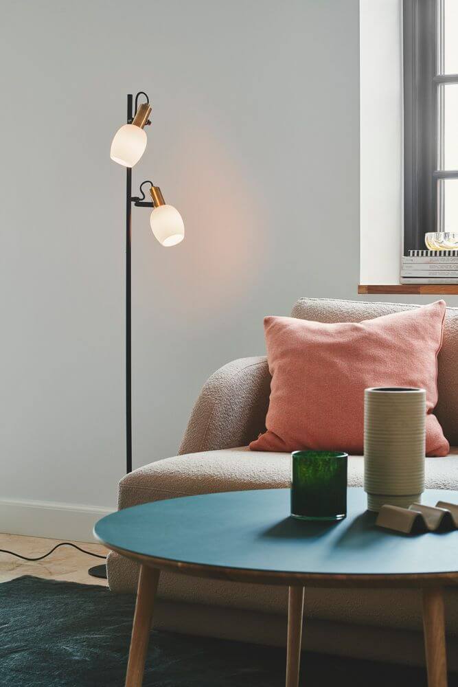 Stehlampe Arild im modernen Raum, mit runden Opalglasschirmen und Messingdetails, dekoriert neben Sofa und Tisch.