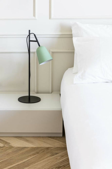 Studio table lamp Ø 11 cm on a bedside table next to a white bed with pillows.