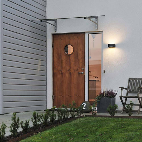 Teramo Außen-Wandleuchte Schwarz, opales Glas neben Eingangstür mit Holz und Garten.