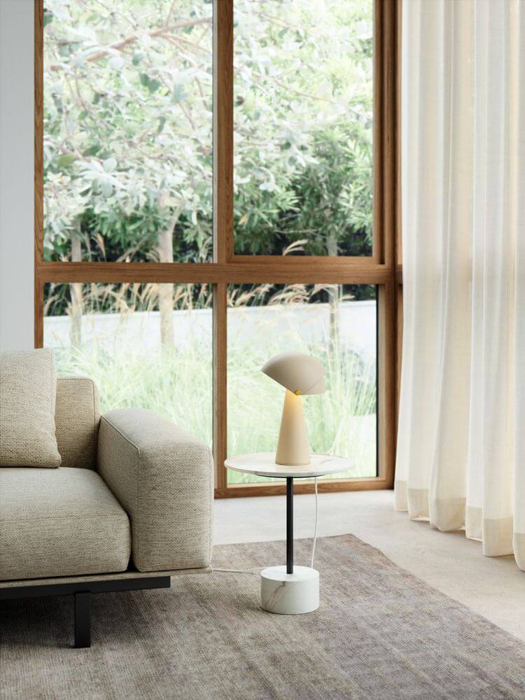 Moderne Wohnzimmer-Einrichtung mit Tischlampe Align neben einem Sofa und großen Fenstern.
