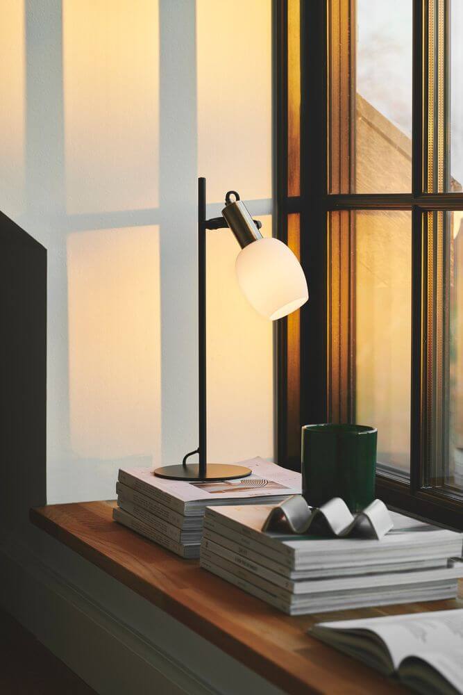 Tischlampe Arild auf einem Tisch mit Bücherstapel und einer grünen Tasse, beleuchtet von Sonnenlicht.
