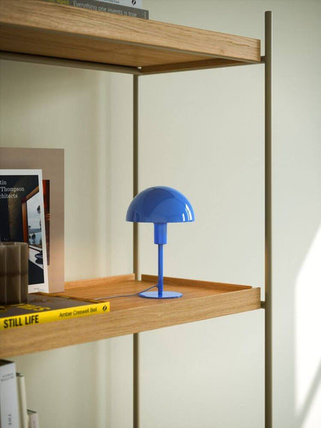 Tischlampe Ellen Mini in Blau auf einem Holzregal, ideal für stilvolle Beleuchtung in kleinen Räumen.