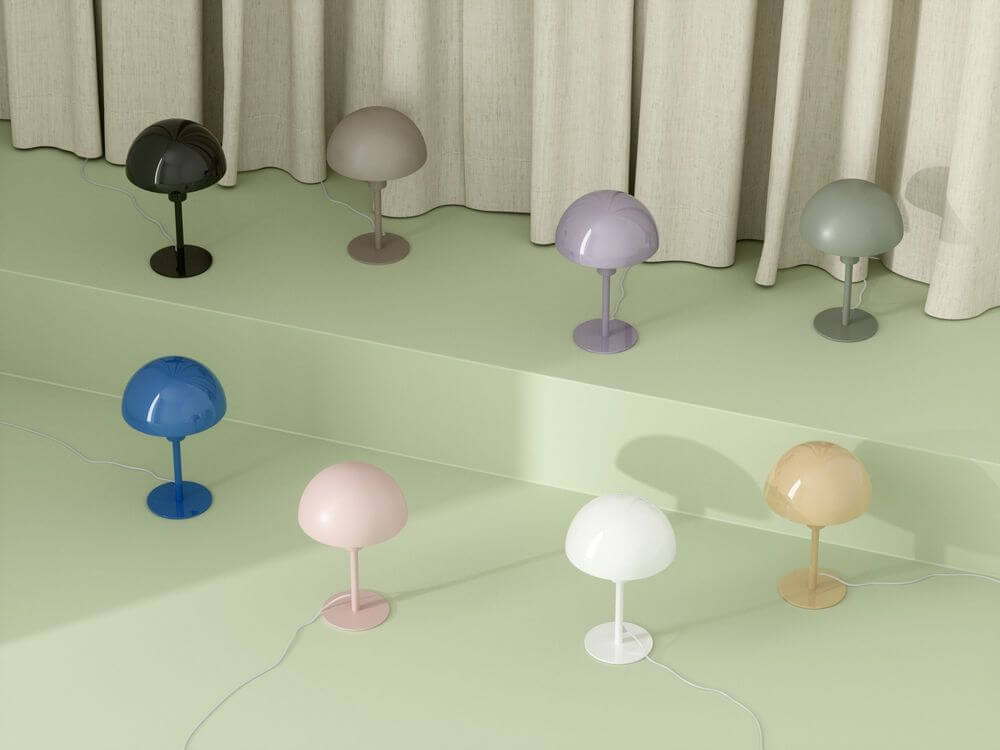 Tischlampe Ellen Mini in verschiedenen Farben auf einer grünlichen Fläche mit einem Hintergrund aus Vorhängen.