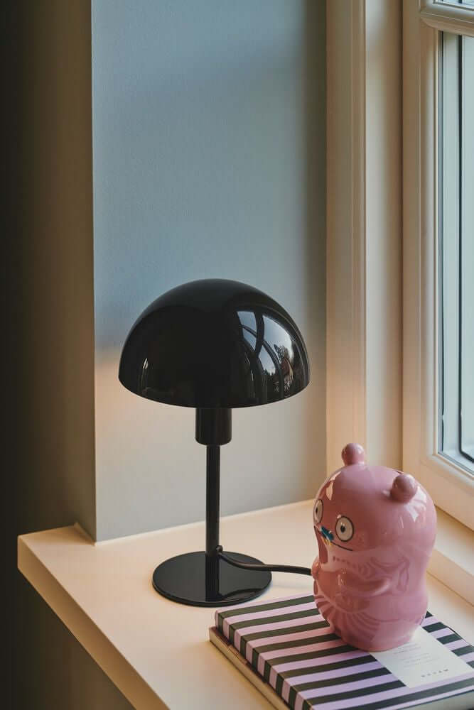 Tischlampe Ellen Mini auf einem Fensterbrett, umgeben von einem vintage rosa Figur und einem Streifenbuch.