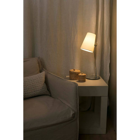 Tischlampe LUPE Chrom, Beige leuchtet neben einem Sofa und dekorativen Körben auf einem Tisch.