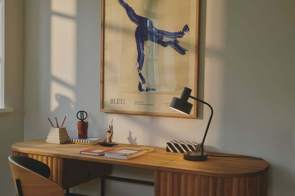 Tischlampe Matis auf einem eleganten Schreibtisch mit modernem Kunstwerk im Hintergrund sowie stilvollen Büromaterialien.