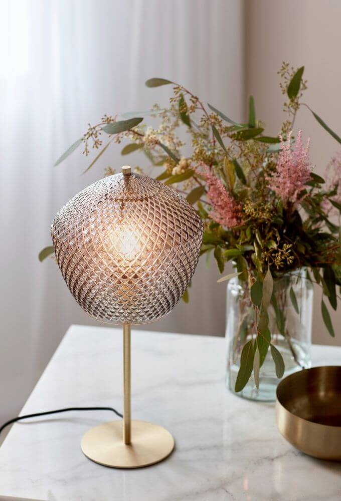 Tischlampe Orbiform mit gebürstetem Messing und gläserner Pentagonform auf einem Tisch, umgeben von Blumen.