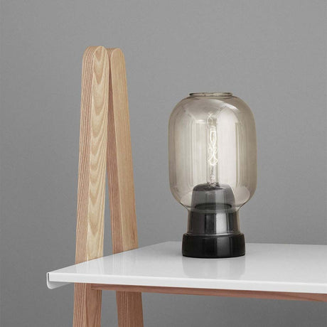 Tischleuchte Amp im Skandi-Look Rauchglas auf einem Tisch mit einem Holzgestell.