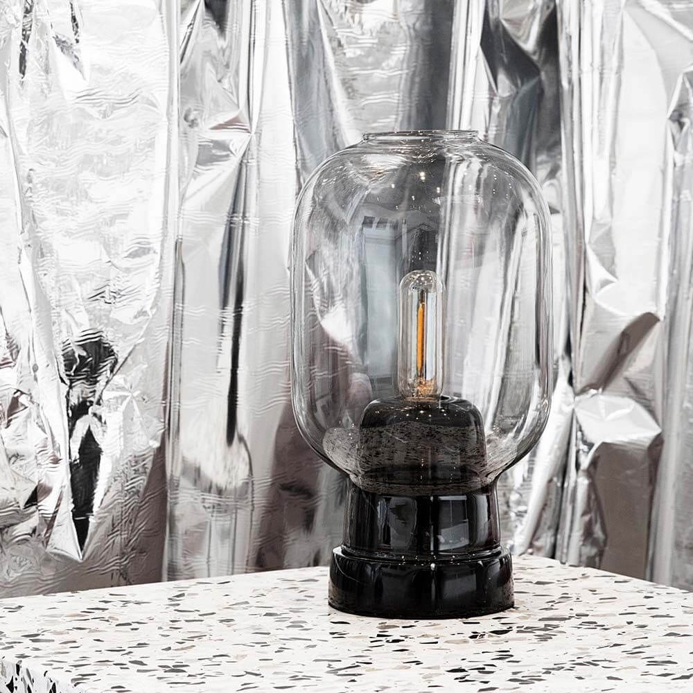 Tischleuchte Amp im Skandi-Look Rauchglas auf einem Tisch mit modernem Design und reflektierendem Hintergrund.