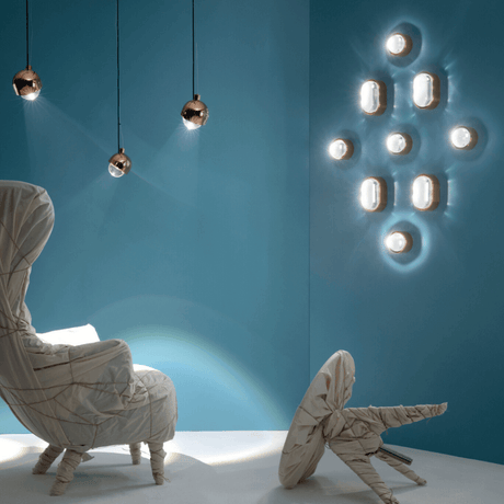 Tom Dixon Spot Wandleuchte mit Glaslinse an blauer Wand, kombiniert mit modernen Pendelleuchten und Sitzmöbeln.