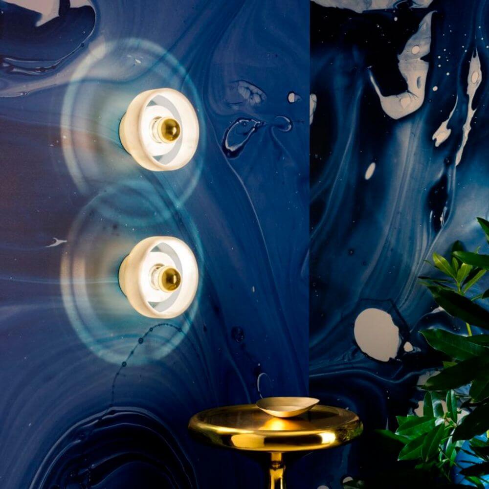 Tom Dixon Stone Wandlampe aus Marmor an einer blauen Wand mit Pflanzen und goldener Tischlampe.
