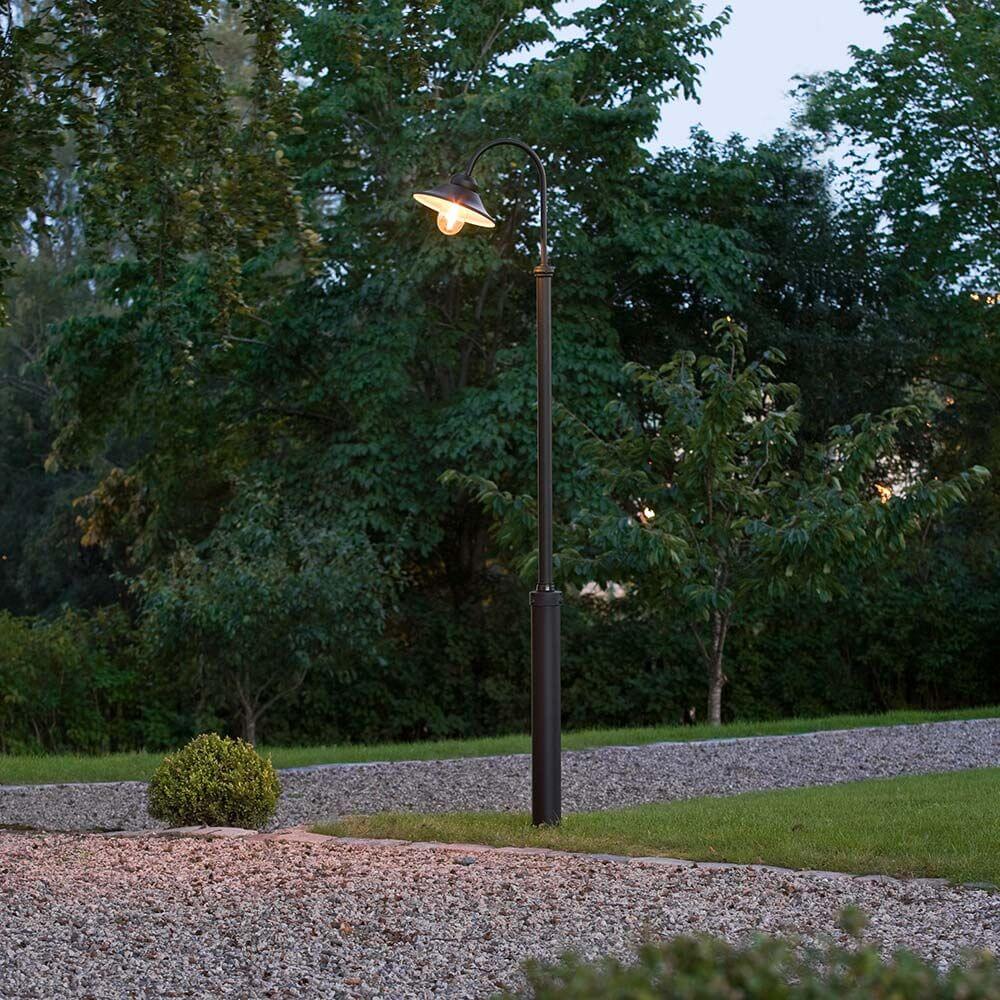 Vega Standleuchte Schwarz, klares Glas im Gartenbereich bei Dämmerung, umgeben von Bäumen und Steinen.