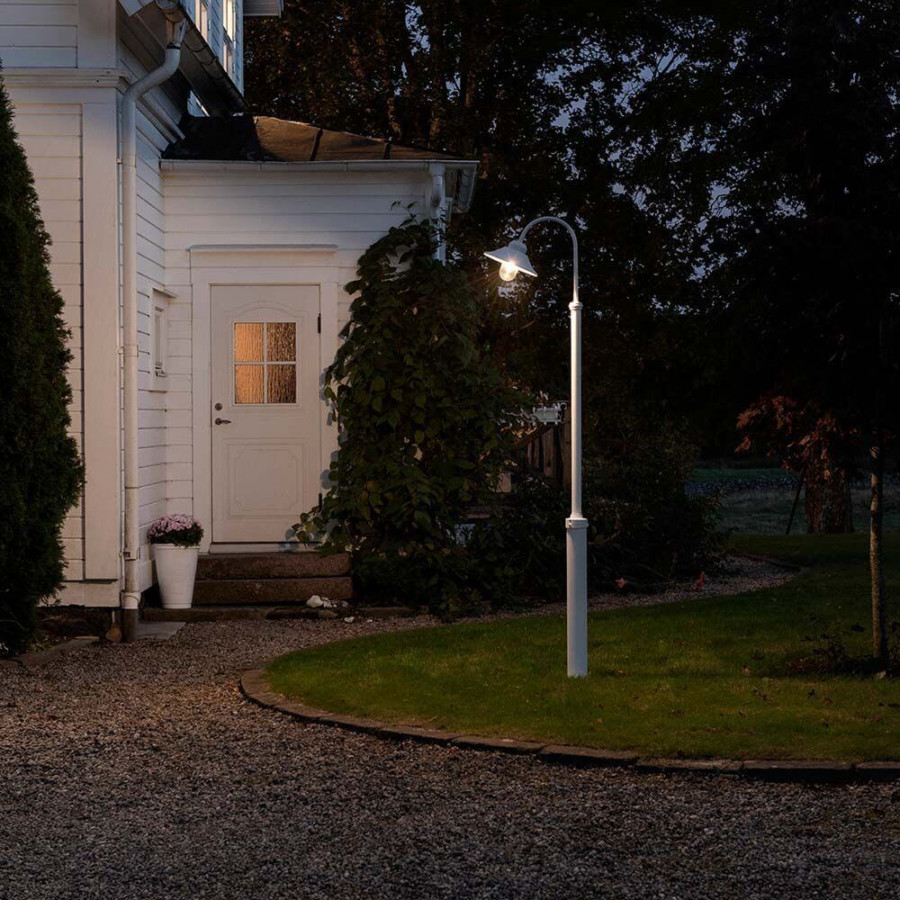 Vega Standleuchte Weiß, klares Glas leuchtet im Garten vor einem weißen Haus bei Nacht.