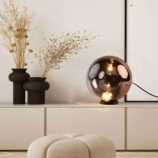Moderne Tischlampe mit runder, bronzefarbener Glaskugel auf einem minimalistischen Sideboard, umgeben von Trockenblumen.