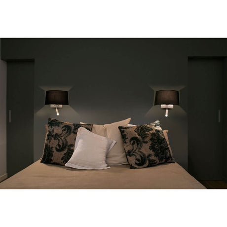 Wandlampe VOLTA mit LED-Leselicht 2700K Nickel, Schwarz neben einem Bett mit dekorativen Kissen.