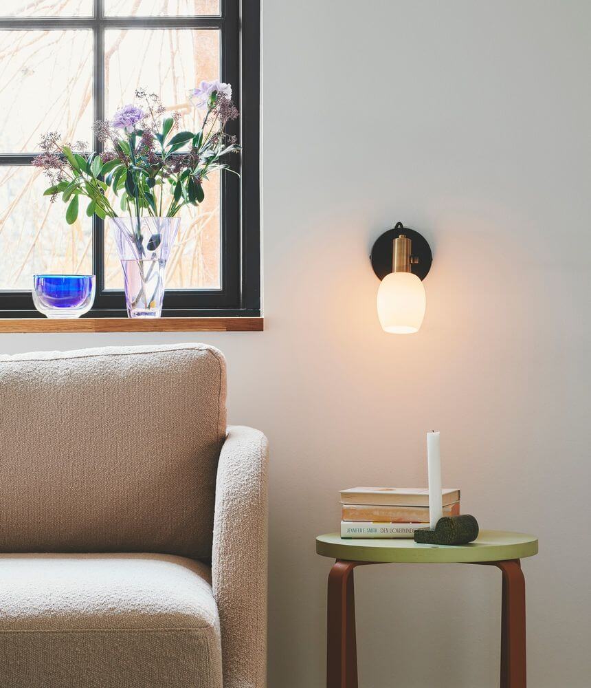 Wandleuchte Arild an einer Wand neben einem gemütlichen Sofa und einem kleinen Beistelltisch mit Büchern.