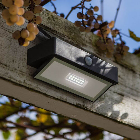 LED-Wandleuchte Arrow mit Solarpanel und Bewegungsmelder an einem Holzbalken montiert, umgeben von Pflanzen.
